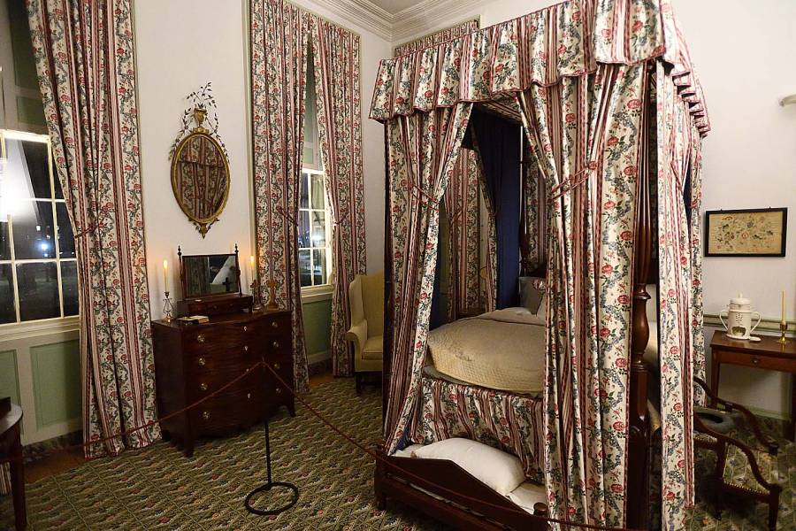 A candlelit bedroom at Homewood Museum includes a four-poster bed with patterned curtains on the posts and a canopy on top. Matching curtains hang from two windows.