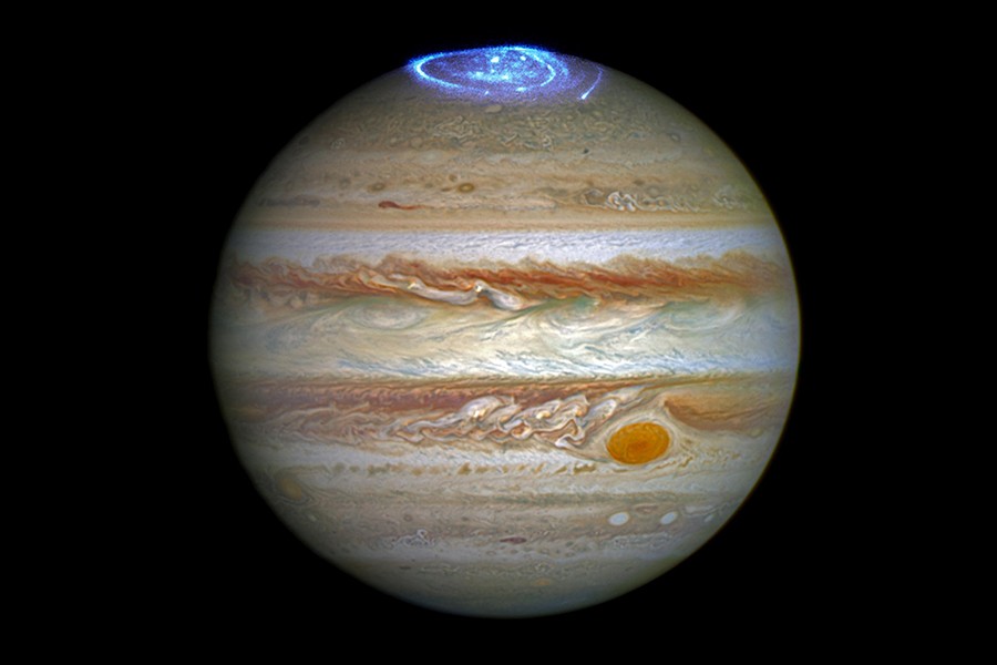 Image depicts a photo of Jupiter, striated with red and gray storm clouds and the iconic Red Dot, topped with an electric blue swirl of color on the north pole representing an aurora