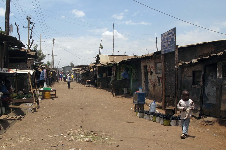A main thoroughfare is a dirt road with corn debris littering the middle. Buildings are assembled with a mix of materials including cardboard, sheet metal, and trash including a FedEx sign