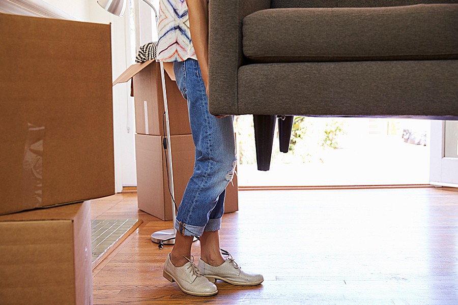 A woman carries one end of a couch into a room filled with moving boxes