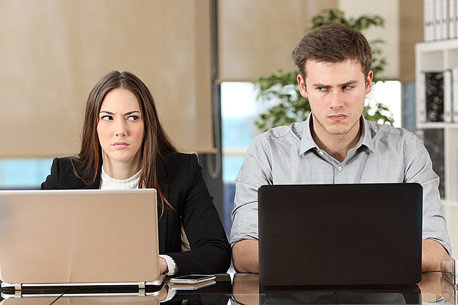 Female and male co-workers glare at each other while working side-by-side on their laptops.