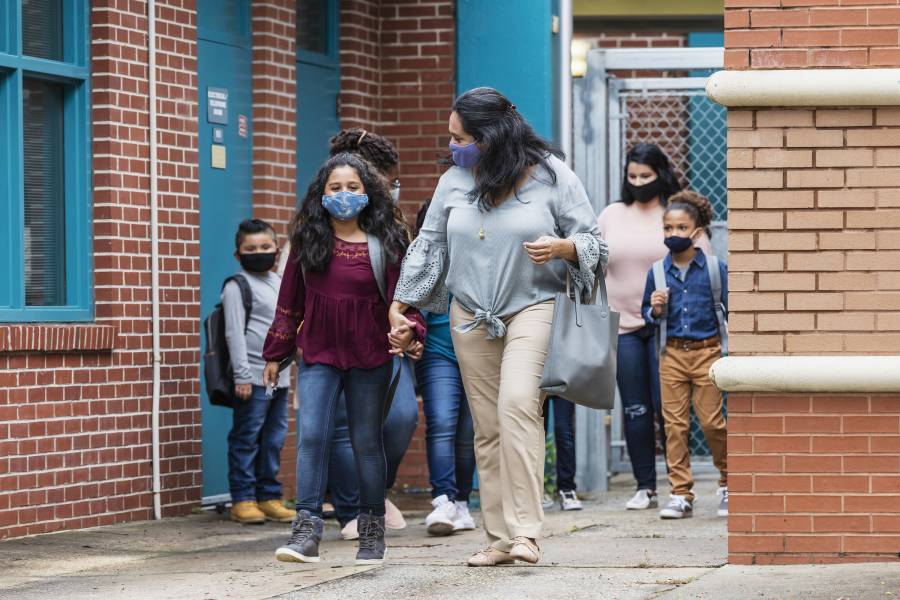 A multi-ethnic group of elementary school children with their mothers, walking outside the school building, wearing protective face masks. 