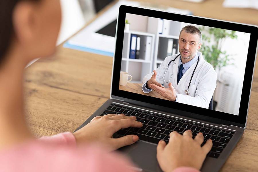 Doctor wearing a white lab coat with a stethoscope around his neck appears on the laptop screen of the patient with whom he's talking