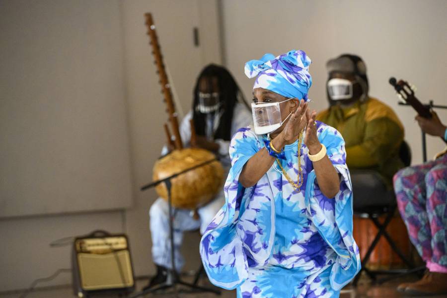 A woman in a tie-dye kaftan and matching head wrap dances while musicians play traditional African folk instruments behind her