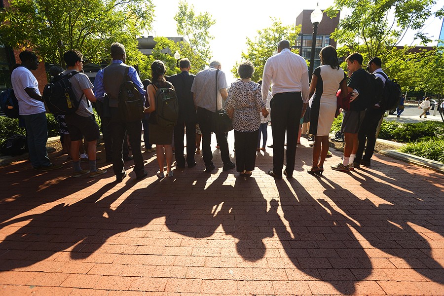 Vigil attendees photographed from behind, holding hands and casting long shadows on a brick walkway