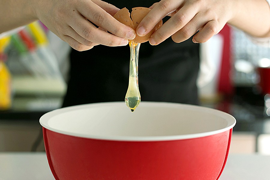 Photo shows two hands cracking a raw egg into a red bowl.