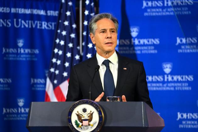 U.S. Secretary of State Antony Blinken stand at a podium in front of a blue backdrop; part of an American flag is visible over his right shoulder