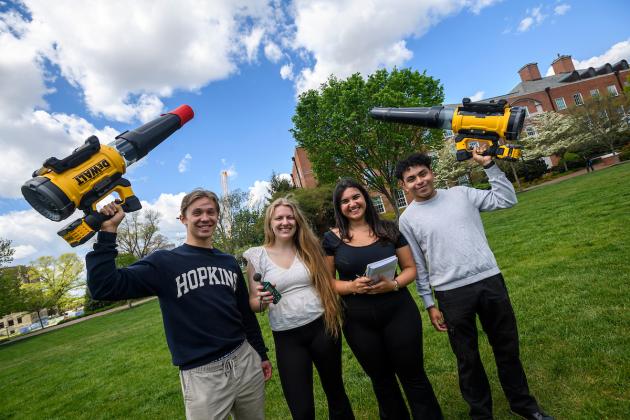 Four Johns Hopkins University students hold up leaf blowers on a quad