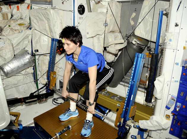 Italian astronaut Samantha Cristoforetti, wearing a blue shirt and black athletic pants, performs a deadlift exercise using the advanced Resistive Exercise Device (aRED) aboard the International Space Station. The aRED machine, equipped with metal bars and vacuum cylinders, provides resistance training to help astronauts maintain muscle and bone strength in microgravity. The surrounding area is filled with white padded walls, equipment, and storage bags, characteristic of the ISS interior.