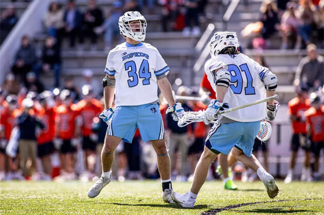 Two men's lacrosse players in white jerseys celebrate a goal