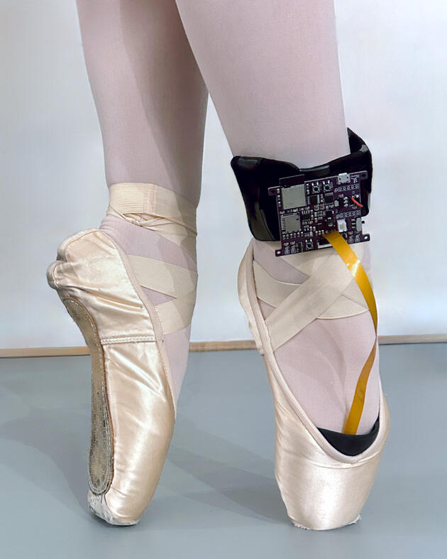 Close-up image of a dancer standing on her toes in ballet pointe shoes; a black control board is wrapped around her right ankle and connected into the toe of the shoe