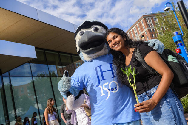 A college student holding a bamboo plant poses for a photo with Jay the Blue Jay in front of the Bloomberg Student Center.