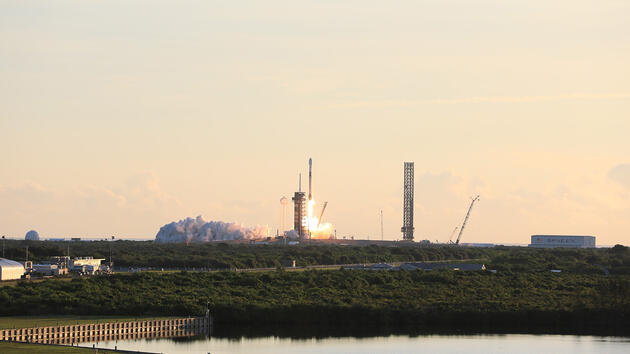 A rocket launches in the distance against a backdrop of a yellow-orange Florida sunrise