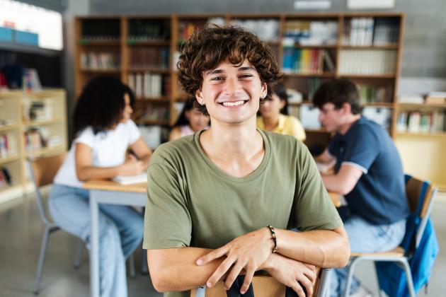 A smiling high school student is in the foreground with his classmates working at tables behind him