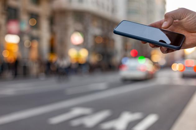 Photograph of a hand holding a cell phone; in the background is a busy street with a taxi lane