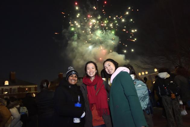 Three college students smile for a picture as fireworks go off in the background.