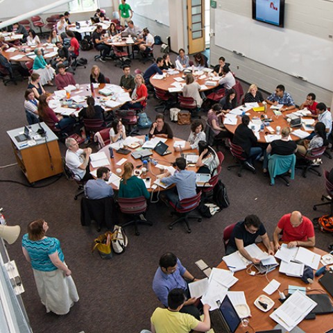 Groups of people sit at round tables with pages and books spread out around them