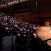 Two people sit on a stage and converse in front of a full auditorium audience